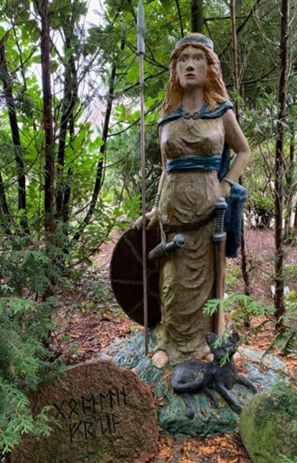 Naive Darstellung einer Freya-Figur. Die Holzfigur trägt Schild und Speer, aber auch betont weibliche Kleidung.