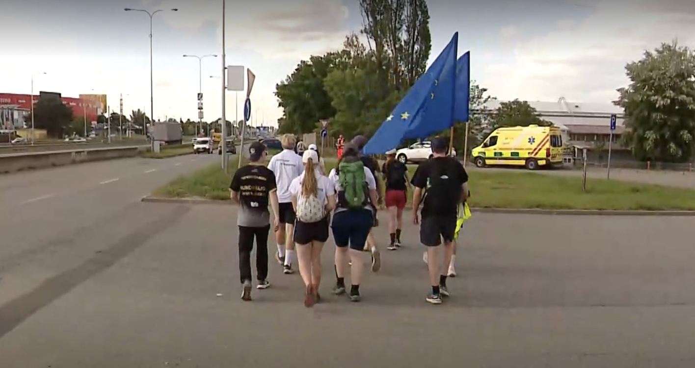 Ein Eindruck vom Gedenkmarsch 2026 im mährischen Brno. Junge Menschen gehen gemeinsam an einer Straße entlang. Rechts lassen zwischen Personen Europa-Fahnen im Wind wehen. 