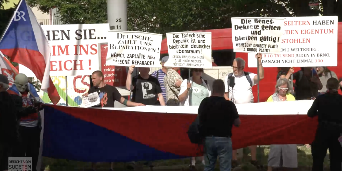 Vor dem Veranstaltungsgelände des Sudetendeutschen Tags in Brno formierte sich Widerstand, der gegen die deutsch-tschechische Kooperation vor allem mit Bezügen auf die NS-Verbrechen und Reparationsforderungen polemisierte. Einige Menschen versammeln sich hinter einen großen Fahne der Tschechischen Republik und halten Schilder hoch.