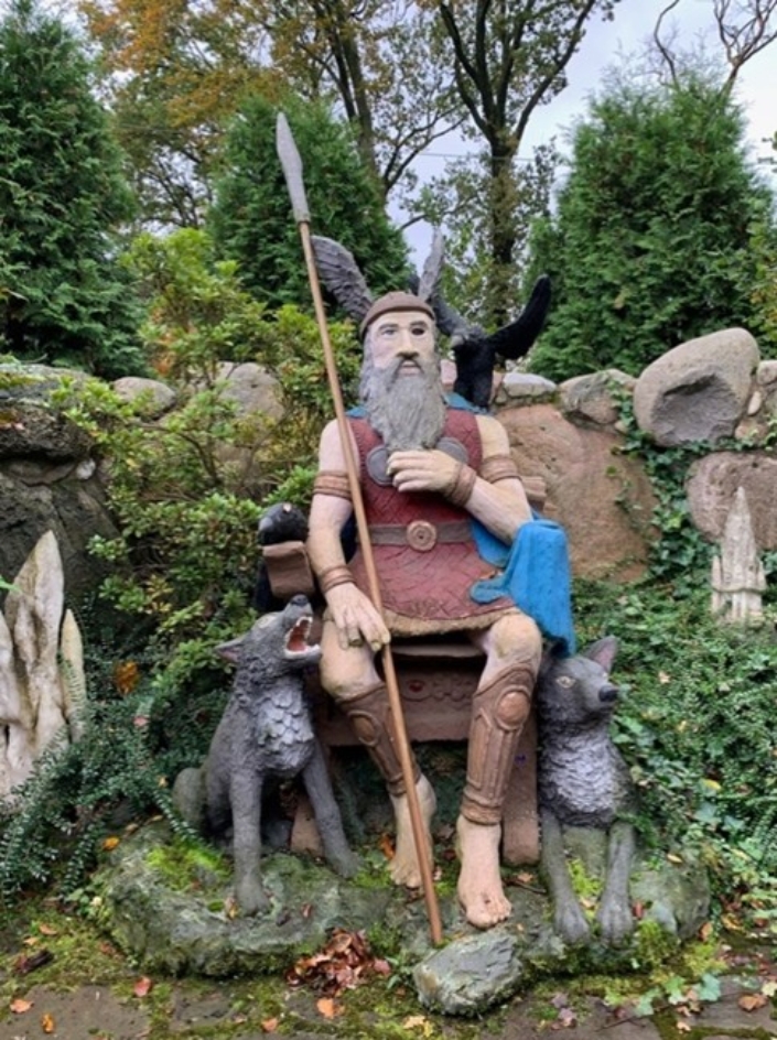 Naive Darstellung einer Odin-Figur aus Holz. Die sitzende Figur hat einen grauen Vollbart, trägt eine Krone sowie einen Speer in der rechten Hand.