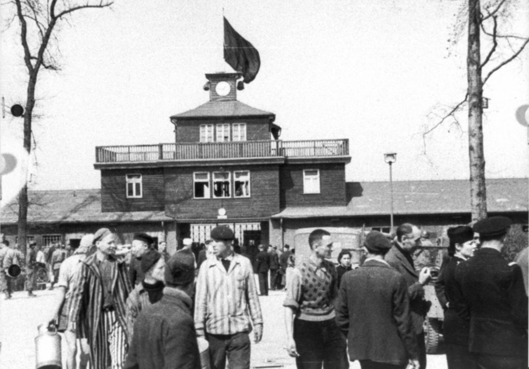 Befreite Häftlinge vor dem Torgebäude, auf dem eine schwarze Fahne gehisst ist. Im Hintergrund Angehörige und Fahrzeuge der U.S. Army