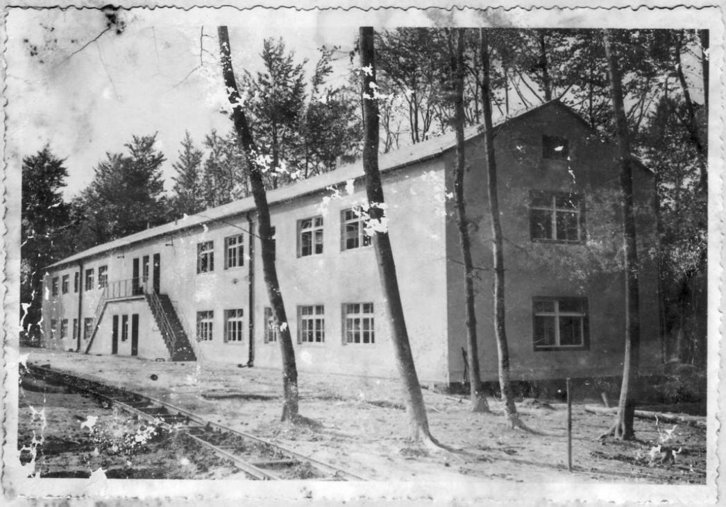 Ein längliches Gebäude mit zwei Stockwerken. An der breiten Seite sind Eingänge für das untere Geschoss, darüber, mit außenliegenden Treppen zu erreichen, ein Eingang für das obere Geschoss. Es ist von Bäumen umgeben. 