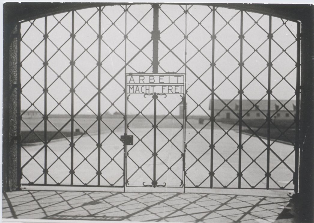 Zu sehen ist ein Eisenes Lagertor. Über der Kleinen Tür in der Mitte steht "Arbeit macht frei" in metallenden Buchstaben.