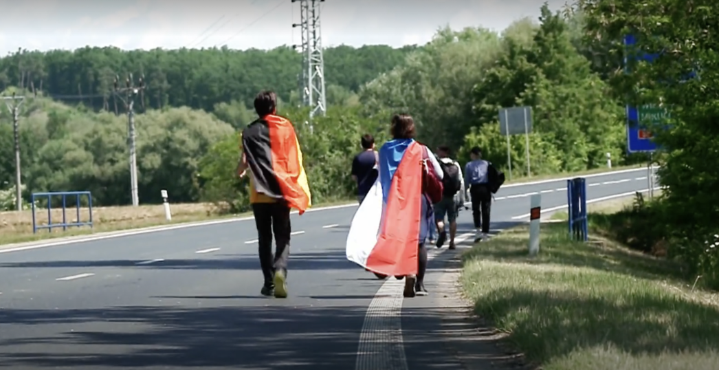 Der mittlerweile alljährlich stattfindende Gedenkmarsch von Brno anlässlich des 80. Jahrestages im Mai 2025. Auf dem Bild sind auf einer Landstraße zwischen Feldern und Wiesen zwei junge Menschen zu sehen: Der linke hat eine Deutschlandfahne über den Rücken hängen, der rechte einen Fahne der Tschechischen Republik. 