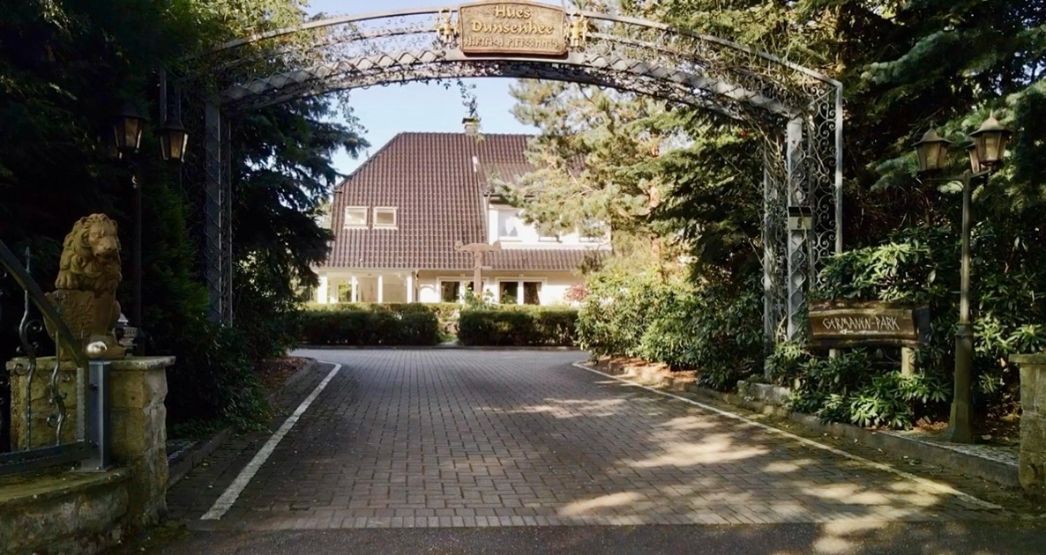 Toreinfahrt zum sogenannten Germanenpark. Links auf einer Mauer sitzt eine mythische Figur, am Ende der Einfahrt ist ein großes Landhaus zu sehen.