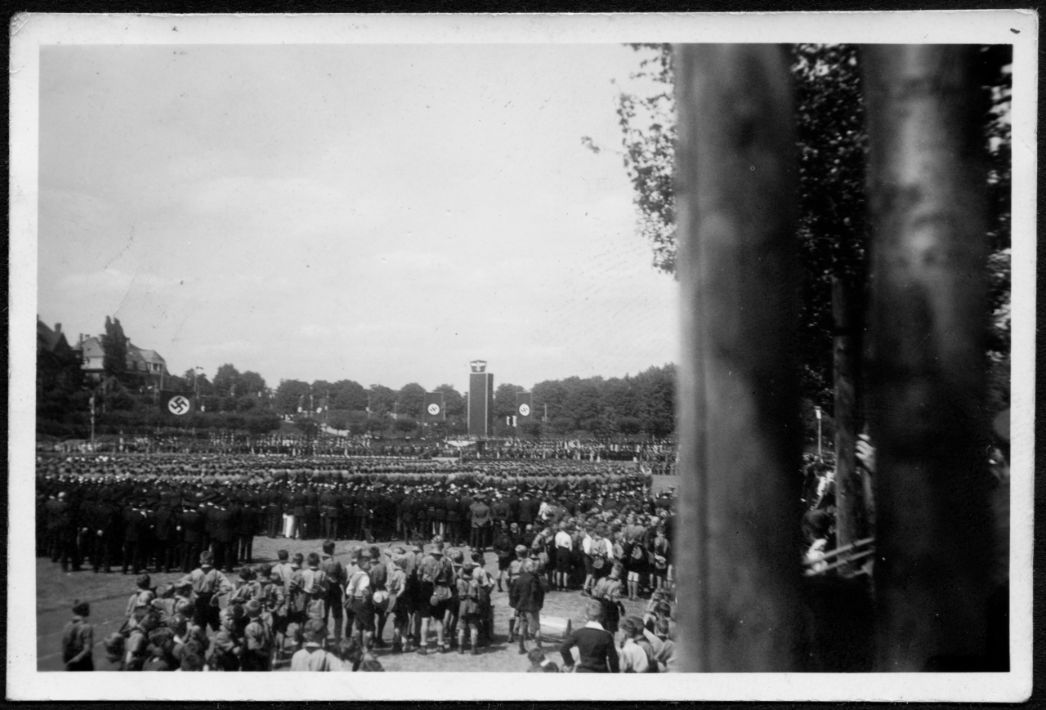 Großkundgebung der Hitlerjugend am 4. August 1935 in Bonn: Tausende uniformierte HJ-Mitglieder versammeln sich auf einem von Bäumen gesäumten Platz; im Hintergrund erhebt sich ein Turm, geschmückt mit großen Hakenkreuzfahnen.