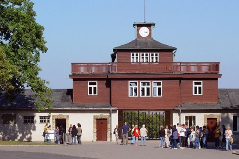 Das Torgebäude. Zwei Flügel, darüber ein kleiner Turm mit einer Uhr an der Spitze. Unter dem Turm das Tor. Davor Gruppen von Besucher:innen