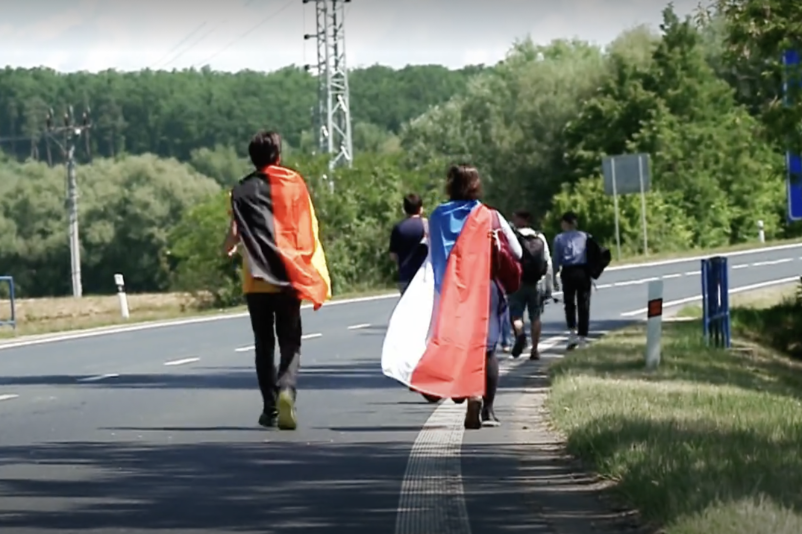 Der mittlerweile alljährlich stattfindende Gedenkmarsch von Brno anlässlich des 80. Jahrestages im Mai 2025. Auf dem Bild sind auf einer Landstraße zwischen Feldern und Wiesen zwei junge Menschen zu sehen: Der linke hat eine Deutschlandfahne über den Rücken hängen, der rechte einen Fahne der Tschechischen Republik. 