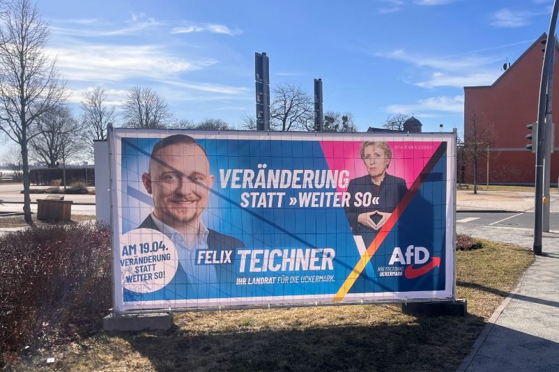 Das Wahlplakat zeigt den Herausforderer Felix Teichner (AfD) sowie die amtierende Landrätin Karina Dörk (CDU) vor einem rosafarbenen Dreieck, das als „Rosa Winkel“ interpretiert wurde. Screenshot aus: AfD-Landratskandidat Teichner löst mit Wahlplakat Kontroverse aus, in: rbb, dort datiert am 3. März 2026, URL: www.rbb24.de/politik/wahl/kommunalwahlen/beitraege-2026/landratswahl-uckermark-doerk-teichner-wahlplakate.html (9. März 2026).